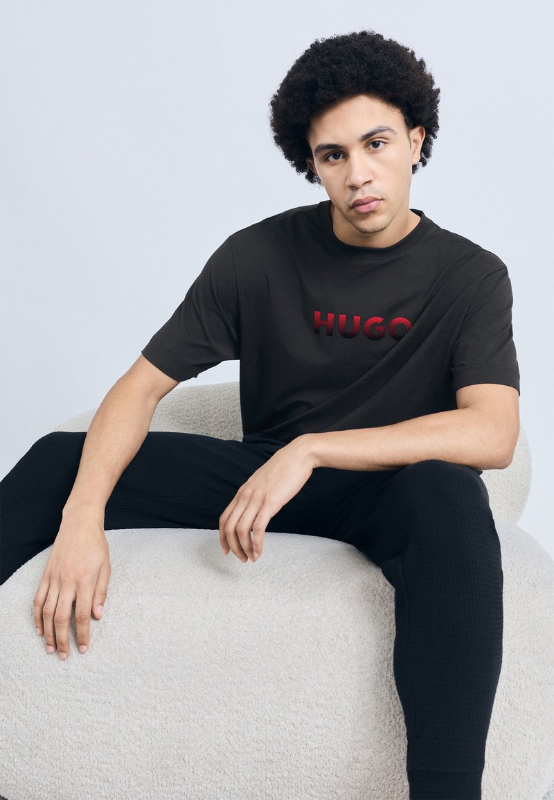 Čierne tričko s krátkym rukávom s vystúpeným červeným logom „HUGO“, nosené s texturovanými čiernymi nohavicami, na neutrálnej pozadí. Mäkké, plyšové sedenie.
