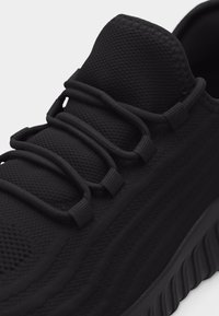 Κοντινή λήψη μαύρου αθλητικού παπουτσιού με υφή υφάσματος στην επάνω επιφάνεια και παχιές κορδόνια περασμένα μέσα από υφασμάτινα θηλύκια στην κορυφή του παπουτσιού.