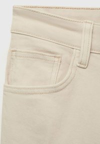 Beige denim fabric with a textured weave, featuring a front pocket outlined with contrasting stitching and a belt loop at the waistband.
