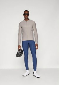 Garām piedurknēm pelēka riteņbraukšanas džersija ar rāvējslēdzēju priekšpusē, kopā ar zilām legingām. Modelis tur melnu ķiveri, valkājot baltas sporta kurpes.