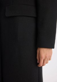 Czarny wełniany płaszcz o gładkiej fakturze, z wyraźną konstrukcją i pojedynczą, wyraźną kieszenią w górnej części rękawa.
