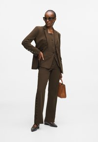 Brauner maßgeschneiderter Anzug mit einer taillierten Blazerjacke, Hose mit geknöpften Seitendetails, kombiniert mit spitzen schwarzen Schuhen und einer braunen Handtasche.