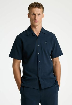 Junger Mann mit lockigem Haar, der ein marineblaues kurzärmliges Hemd mit Knöpfen und passende Hose trägt, Hände in den Taschen, neutraler Hintergrund.