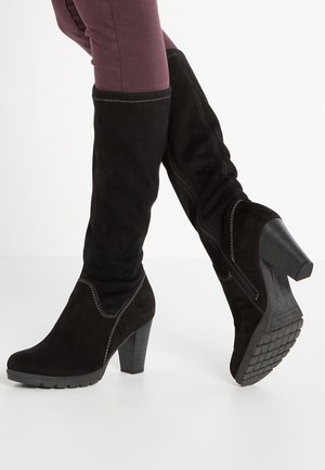 Person wearing black suede knee-high heeled boots with stitching details and textured sole, paired with purple fitted pants.