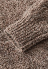 Tessuto di un maglione lavorato a maglia marrone con una texture pelosa, caratterizzato da dettagli a coste sui polsini. Il materiale appare morbido e caldo, con leggeri filamenti visibili.
