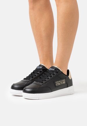 Baskets basses en cuir noir avec semelles blanches et talons ornés d'accents dorés, de la marque "Versace Jeans Couture", portées pieds nus sur un fond uni.