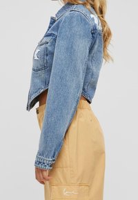Mujer vestida con una chaqueta de mezclilla azul corta con parches blancos y pantalones de cintura alta beige con un bolsillo lateral y un logo blanco.