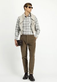 Model ubrany w koszulę w kratkę z guzikami, beżowy sweter dzianinowy narzucony na ramiona, brązowe spodnie kordowe oraz czarne buty, trzymający ciemno-brązową kopertówkę.