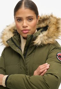 Olijfgroene pufferjack met een faux fur-voering aan de capuchon, rits- en drukknoopsluitingen, en een logo-patch op de linkerarm. Gelaagde gebreide trui eronder.