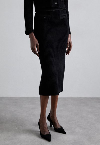Schwarzer Strick-Bleistiftrock mit strukturiertem Stoff, taillierter Silhouette und zwei vorderen Taschen. Kombiniert mit schwarzen spitzen High Heels.