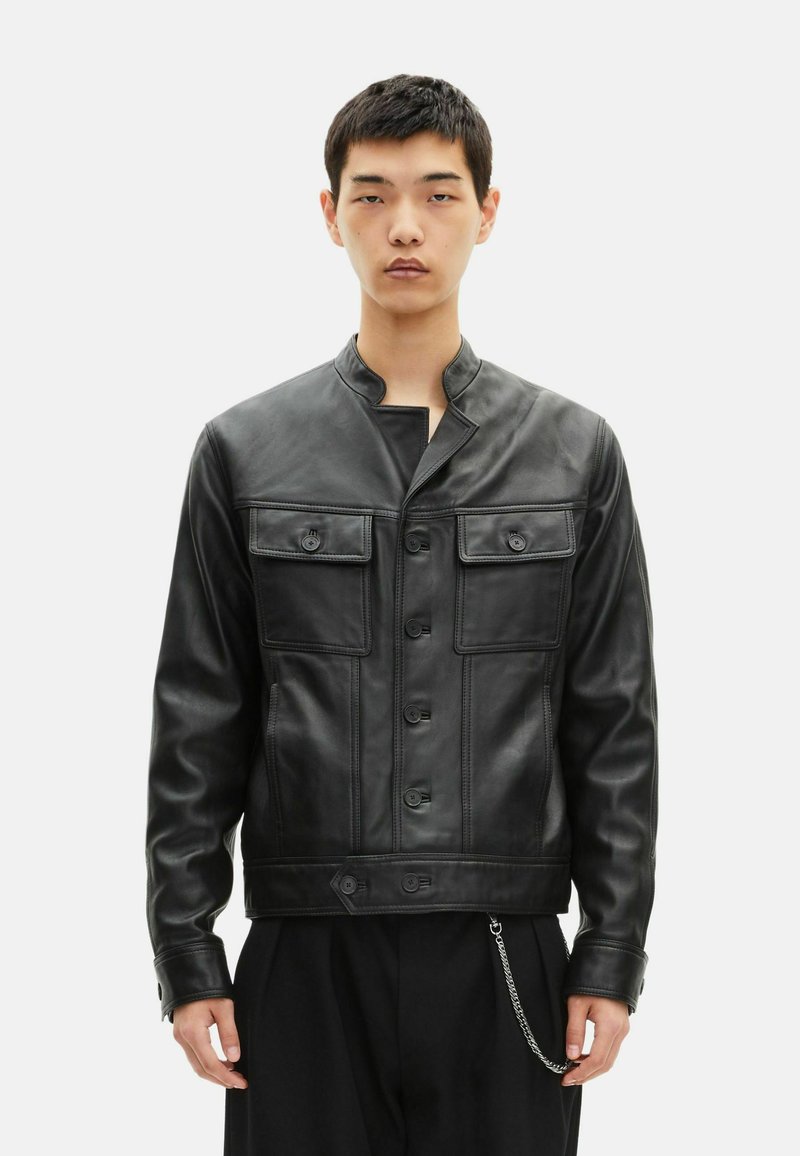 Chaqueta de cuero negra con cuatro bolsillos abotonados, cuello mao y cierre frontal de botones. Presenta un detalle de cadena en el dobladillo.