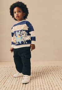 Felpa a righe navy e bianche con personaggi dei cartoni animati, abbinata a pantaloni cargo neri e sneakers bianche con chiusura in velcro. Sfondo di tappeto strutturato.