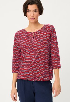 Woman with short dark hair wearing a red patterned blouse with 3/4 sleeves and navy blue pants, standing against a plain light background.