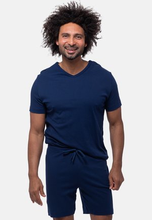 Lachende man met krullend haar, gekleed in een navyblauw shirt met korte mouwen en bijpassende korte broek, tegen een effen achtergrond.