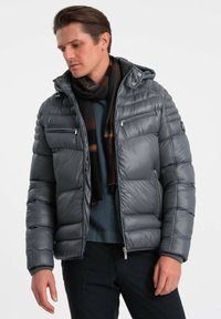 Giacca piumino grigia con cappuccio con zip, texture trapuntata, due tasche frontali con zip e una sciarpa calda. Indossata sopra un maglione blu.