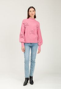 Donna che indossa un maglione rosa con dettagli a rosa testurizzati, jeans azzurri chiari e stivaletti neri alla caviglia, in piedi di fronte a uno sfondo chiaro e semplice.