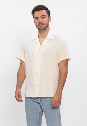 Mann trägt ein cremefarbenes Kurzarmhemd und hellblaue Jeans, steht vor einem schlichten weißen Hintergrund.