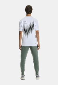 Witte t-shirt met een zwart grafisch ontwerp op de achterkant, gecombineerd met groene sweatpants. T-shirt heeft een ronde hals en korte mouwen.