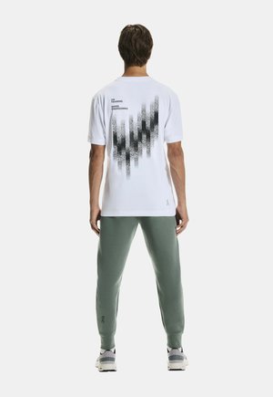 Hvid t-shirt med et sort grafisk design på ryggen, parret med grønne sweatpants. T-shirten har rund halsudskæring og korte ærmer.