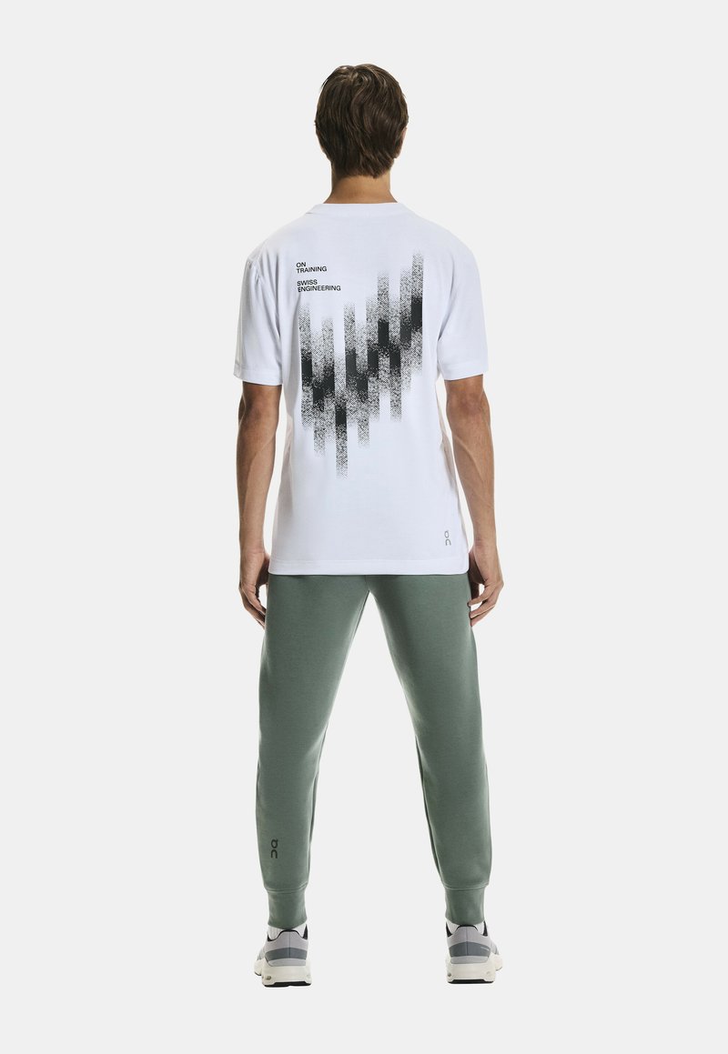 Witte t-shirt met een zwart grafisch ontwerp op de achterkant, gecombineerd met groene sweatpants. T-shirt heeft een ronde hals en korte mouwen.