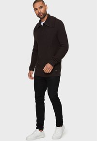 Brauner gerippter Pullover mit hohem Kragen und halbem Reißverschluss, kombiniert mit schwarzen Hosen und weißen Sneakers. Fokus auf warme Texturen und lässige Passform.