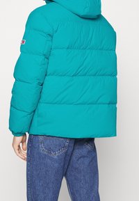 Teal puffjacka med huva, med horisontellt quiltade sektioner och elastiska ärmslut. Distinkt logotyp patch på vänster ärm.