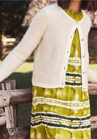 Cremfarbener, fluffiger Cardigan mit Knopfleiste, kombiniert mit einem grün-weißen, gemusterten Kleid, das vertikale Streifen und eine taillierte Taille hat.
