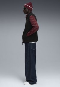 Schwarze Fleece-Weste über einem burgunderfarbenen gestreiften Langarmshirt, kombiniert mit weit geschnittenen dunkelblauen Jeans und weißen Turnschuhen. Eine rote Strickmütze und Sonnenbrille werden getragen.