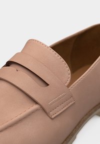 Mocassin rose clair avec une finition lisse, une bride décorative, des coutures visibles et une semelle beige sur fond blanc.