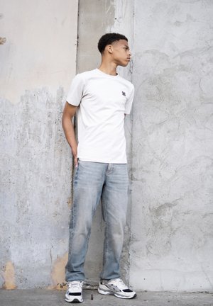 Hvid kortærmet t-shirt med et lille logo, lys blå slidte jeans og hvide sneakers med sorte accenter, lænet op ad en tekstureret væg.