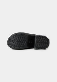 Semelle de chaussure noire avec des motifs de profondeur et une petite partie de matériau blanc au talon, montrée sur un fond gris.