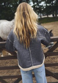 Liela, tumši pelēka jaka ar kontrastējošu beigu šermanu apdari un šuvju detaļām, savienota ar gaiši ziliem džinsiem.