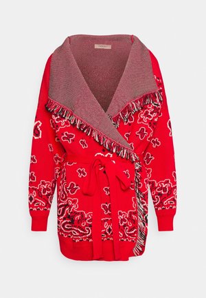 Cardigan en maille rouge à motifs avec col châle à franges et ceinture nouée assortie à la taille sur fond blanc.