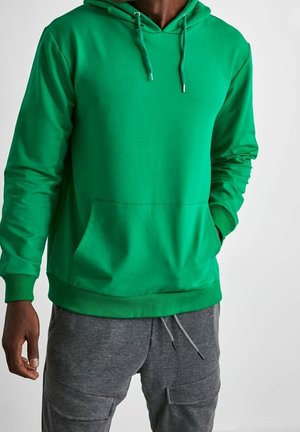 Uomo con felpa verde acceso con tasca frontale e coulisse, abbinata a pantaloni della tuta grigi con tasche cargo.