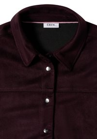 Veste en daim violet foncé avec un col et trois boutons-pression argentés sur le devant, avec une étiquette blanche à l'intérieur du col.