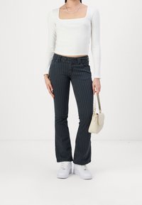 Mujer que lleva una blusa corta de manga larga blanca, pantalones bootcut de raya diplomática oscuros, zapatillas blancas, sosteniendo un pequeño bolso beige a su lado.