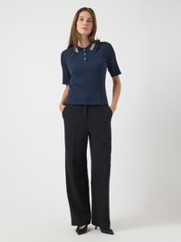 Femme portant un polo à manches courtes en côtes bleu marine avec un col à rayures blanches et un pantalon large noir, se tenant les mains dans les poches.