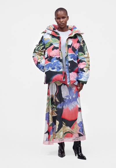 Femme portant une doudoune colorée à motifs et une longue jupe assortie avec des bottines noires à talons, debout devant un fond uni.