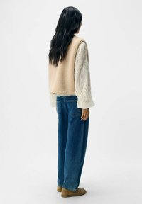 Beige fleece vest over een textuurwit langemouw t-shirt, gecombineerd met losse blauwe denim jeans en tan schoenen, gezien van achteren.