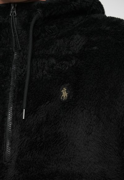 Hanorac cu fermoar negru din fleece, cu o textură moale, având un mic logo brodat în aur. Fermoarul și tab-urile de tragere negre completează designul.