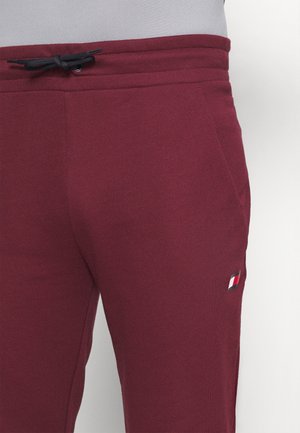 Pantalones deportivos burdeos hechos de tela suave. Cuentan con una cintura con cordón, bolsillos laterales y un pequeño parche con logo en rojo, blanco y azul.