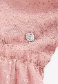 Tissu en maille rose avec de petits points dorés et une étiquette ronde en argent cousue avec du fil rose, gravée des lettres "M*V".