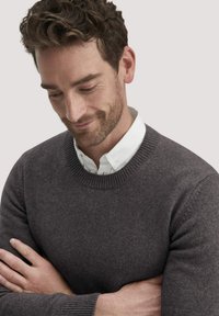 Dunkelgrauer gestrickter Pullover mit rundem Ausschnitt, langen Ärmeln und gerippten Bündchen, über einem weißen Hemd mit Kragen getragen. Weiche Textur.