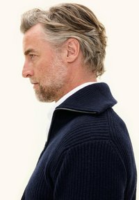 Uomo di mezza età con capelli e barba grigi, indossa un maglione lavorato a maglia blu navy con zip e colletto alto, rivolto verso sinistra su uno sfondo semplice.