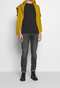 Camisa corta de manga en color navy con lunares amarillos, combinada con un cárdigan texturizado mostaza que cae sobre los hombros, jeans grises y botas negras.