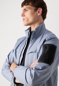 Light blue waterproof jacket with a high collar, zip closure, and a black zippered pocket on the left arm; smooth texture, sporty design.