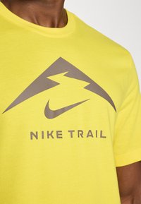 Erkki helekollane puuvillane T-särk, millel on hall kolmnurga logo ja all "NIKE TRAIL" tekst. Ümar kaelus ja lühikesed varrukad.