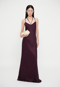 Vestido violeta até ao chão, com alças finas, um decote profundo e uma textura suave. O modelo segura uma carteira de mão creme.