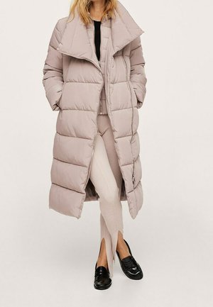 Kvinde iført lang beige dunjakke, beige ribstrikkede bukser med forreste slidser, sorte loafers og sort top, stående mod en ensartet lys baggrund.