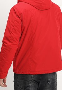 Rød foret jakke med hætte, der har en glat tekstur, elastiske manchetter og en kort pasform. Simpelt design uden synlige metaldele eller mønstre.